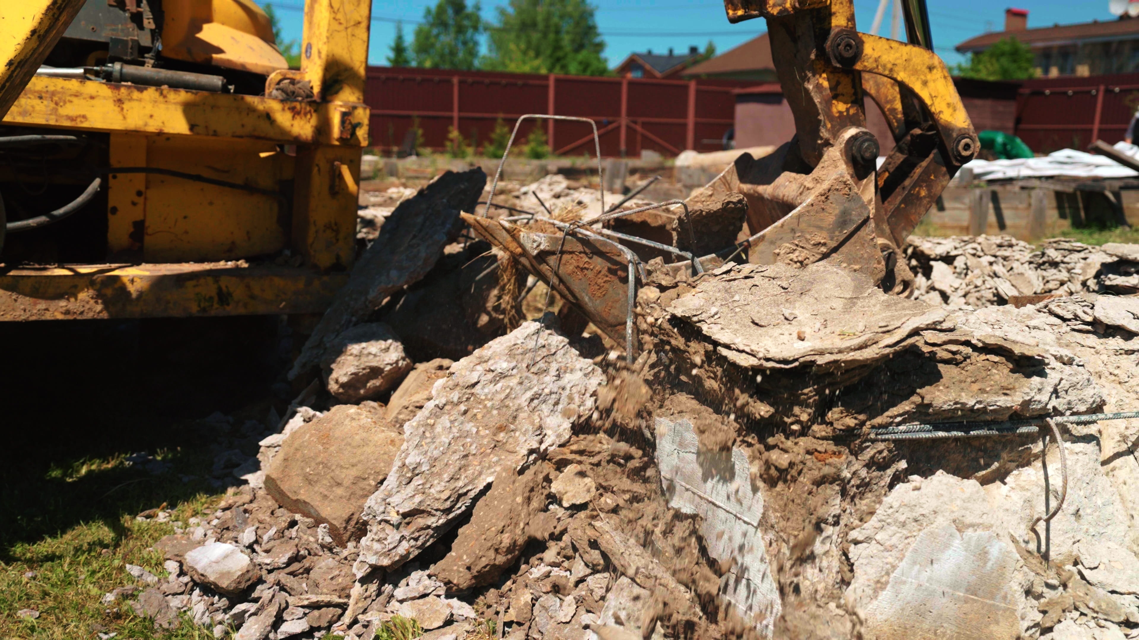 A concrete worker uses both hands to load broken concrete pieces into a wheelbarrow beside a cleared section of slab, with additional debris gathered nearby